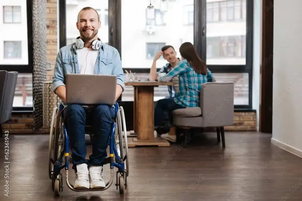 Ein Mann im Rollstuhl arbeitet mit einem Laptop, im Hintergrund unterhalten sich zwei Personen.